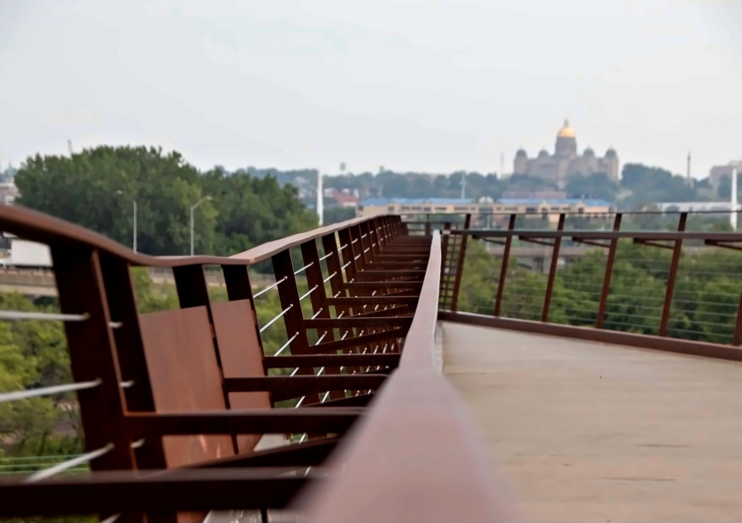 Steel railing of a pedestrian bridge leading toward the Iowa State Capitol and green trees